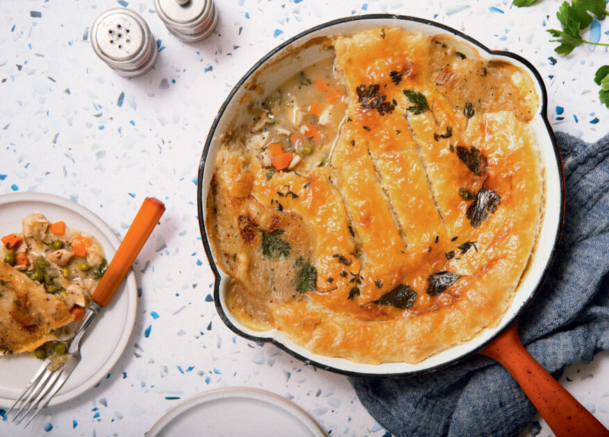 A skillet chicken pot pie with golden crust, some served on a plate beside it, on a white speckled tabletop.