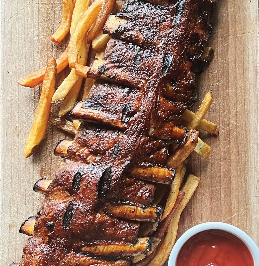 Barbecued rack of ribs with a side of French fries on a wooden board, served with a small dish of ketchup.