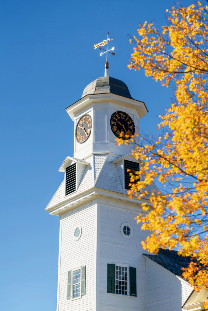 White clock tower with a weather vane against a clear blue sky, partially framed by a tree with bright yellow autumn leaves.