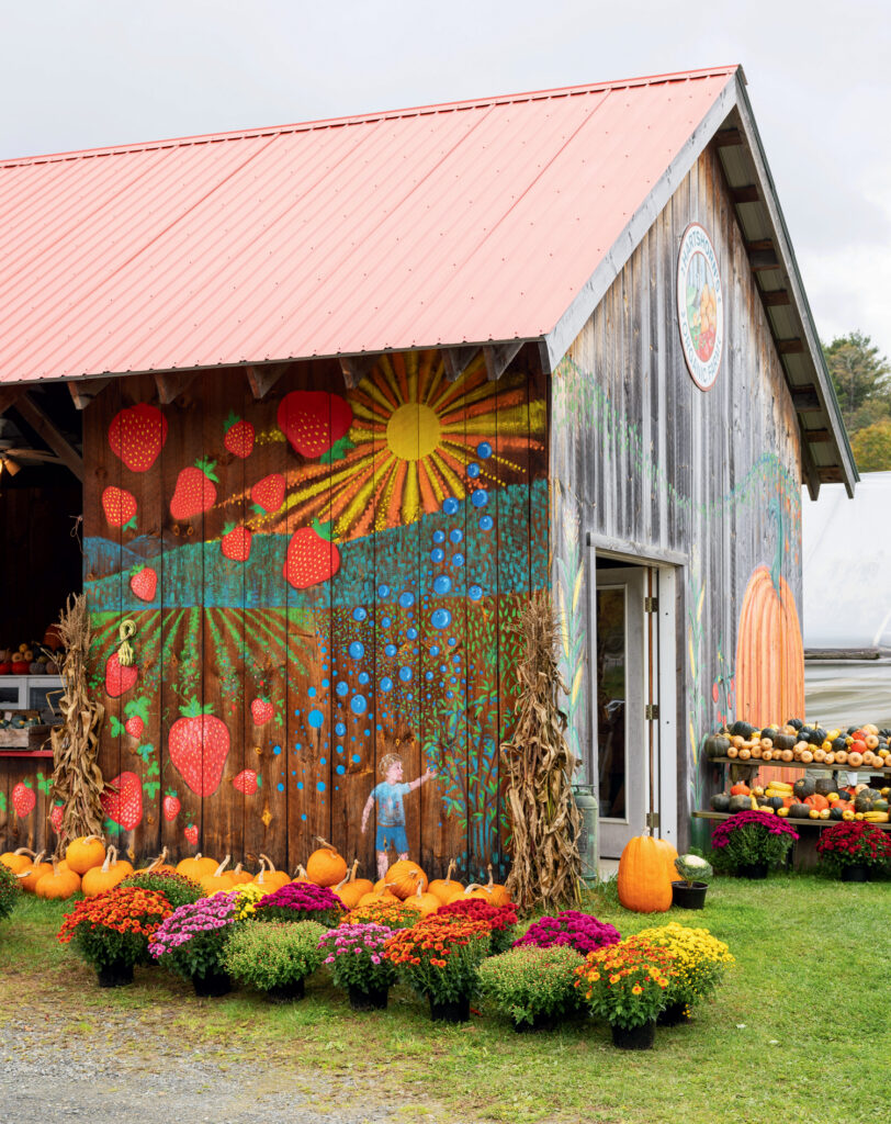 A wooden barn with colorful fruit and sun murals, surrounded by pumpkins, potted flowers, and autumn decorations, on a grassy area under a cloudy sky.