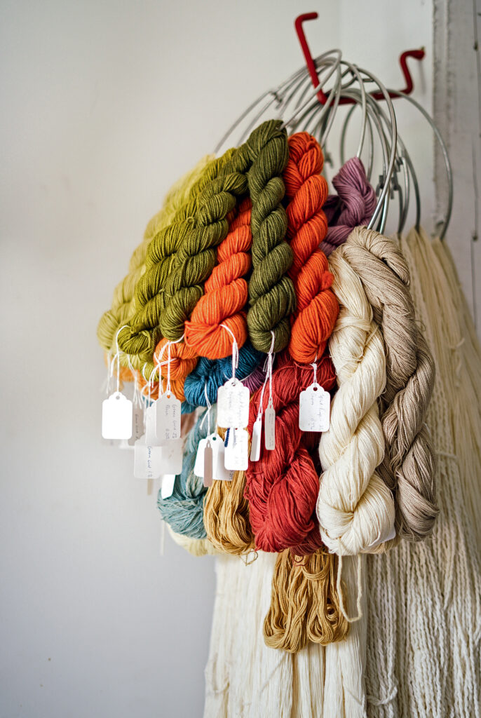 Swans Island Company. Several skeins of colored yarn with tags hang from metal rings against a white wall, arranged in shades of green, orange, red, blue, beige, and purple.