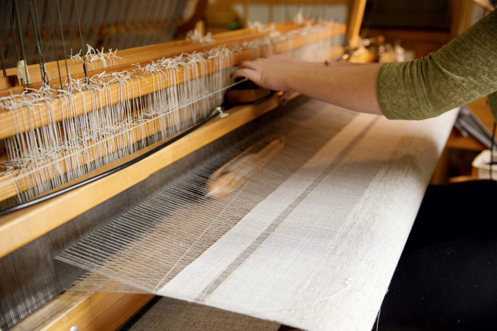 A person operates a wooden loom, weaving fabric with thin, light-colored threads in a workshop setting.