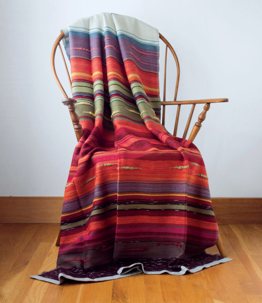 Swans Island Company. A wooden chair with a colorful striped blanket draped over it, placed on a hardwood floor against a plain white wall.