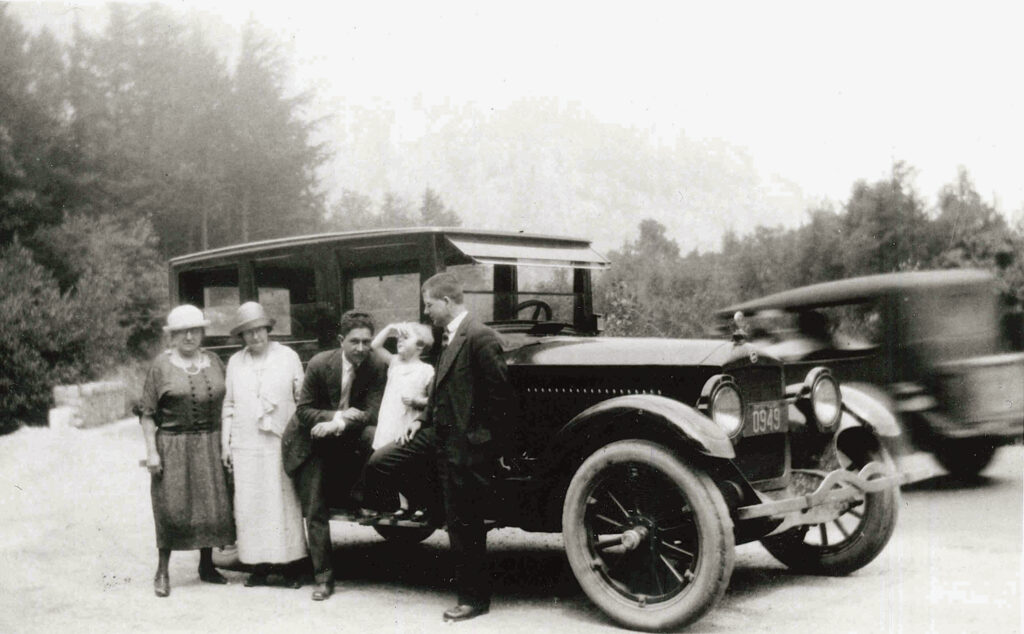 Five people, including a small child, pose beside a vintage car on a road, with trees and another moving car in the background.