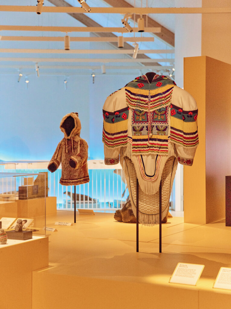 Traditional Indigenous garments are displayed on stands in a well-lit museum exhibit, including a beaded jacket and a fur-lined parka.