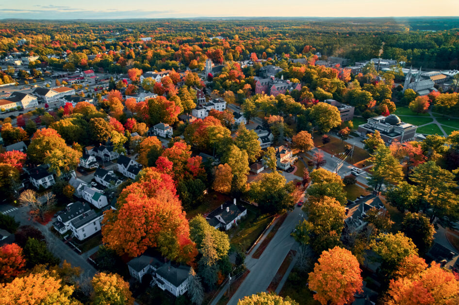 Aerial view of a small town with houses and buildings surrounded by trees displaying fall foliage in shades of red, orange, and yellow.