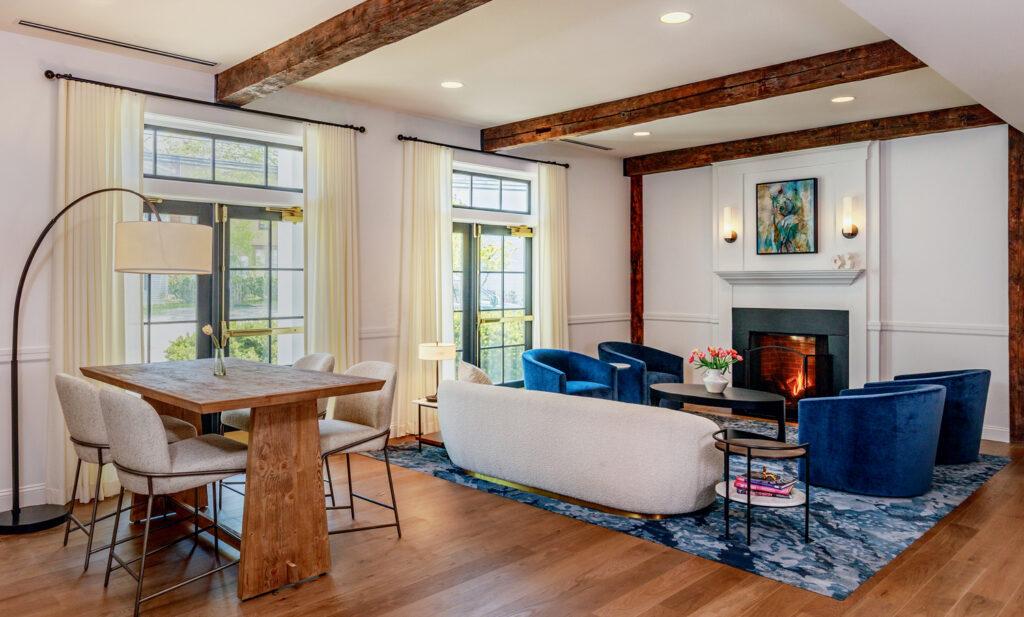 A modern living room with wood beams, a dining table, velvet blue chairs, a white sofa, a fireplace, large windows, and hardwood floors.