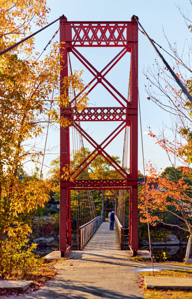 A person walks across a red suspension footbridge surrounded by autumn trees with orange and yellow leaves.