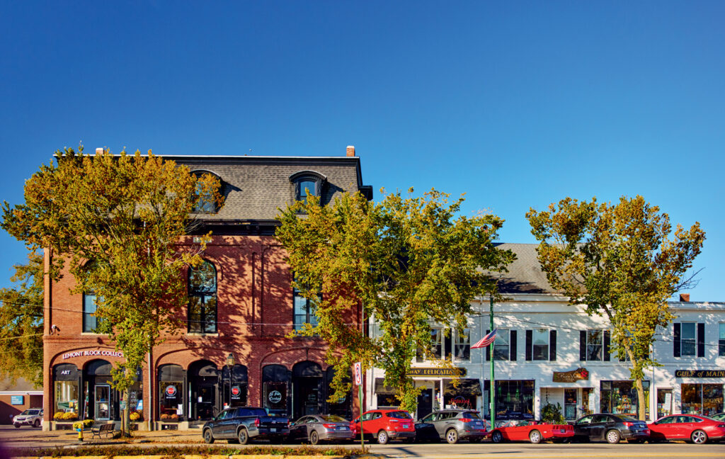 A row of storefronts with brick and white facades, trees lining the sidewalk, and several parked cars on a sunny day under a clear blue sky.