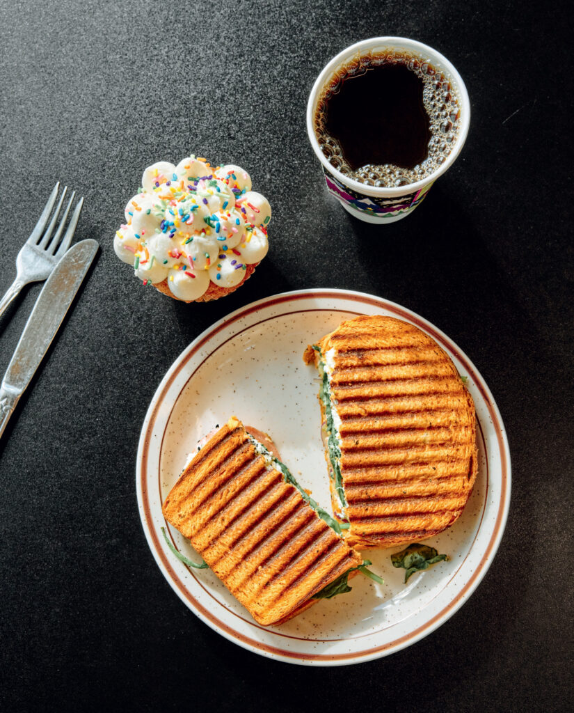 A grilled panini sandwich sits on a plate next to a cupcake with sprinkles and a cup of black coffee. A fork and knife are on the side.