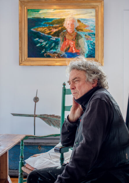 An older man sits pensively in a room with a framed portrait of a woman in a landscape hanging on the wall behind him.