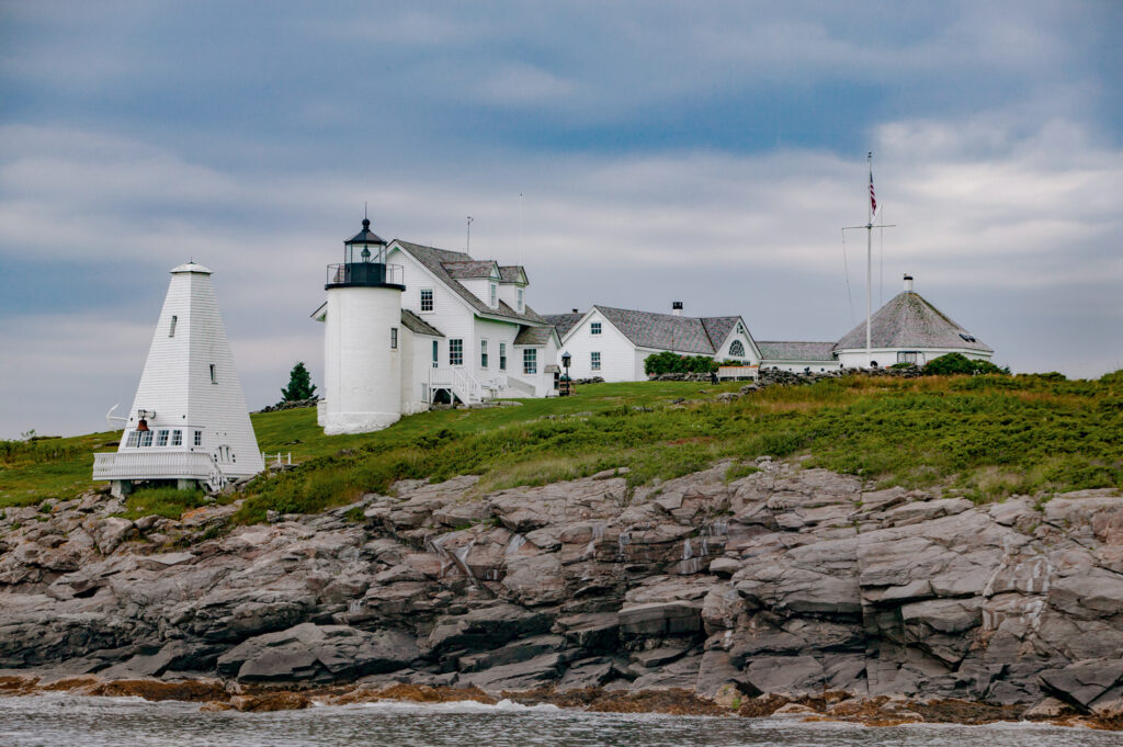 A white lighthouse and adjacent buildings sit on a rocky, grassy coastline under a cloudy sky.