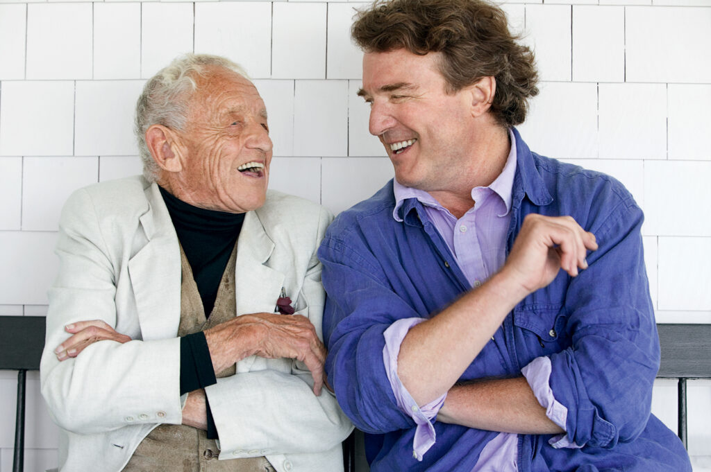 Two men sitting on a bench, smiling and laughing together, with a white tiled wall in the background.