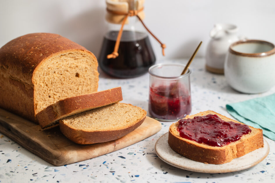 A loaf of sliced Anadama bread on a cutting board, a slice with red jam on a plate, a jar of jam, a mug, a small pitcher, and a carafe of coffee on a countertop.
