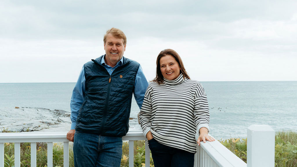 Two people stand side by side, smiling on a deck overlooking the ocean with cloudy skies and rocky shoreline in the background.