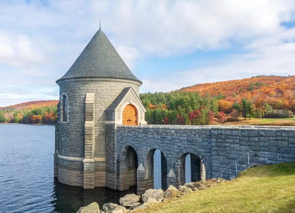 6 Scenic Spots to See New England Fall Foliage. Saville Dam. Stone tower on lake with arched bridge, surrounded by autumn trees and hills under a partly cloudy sky.