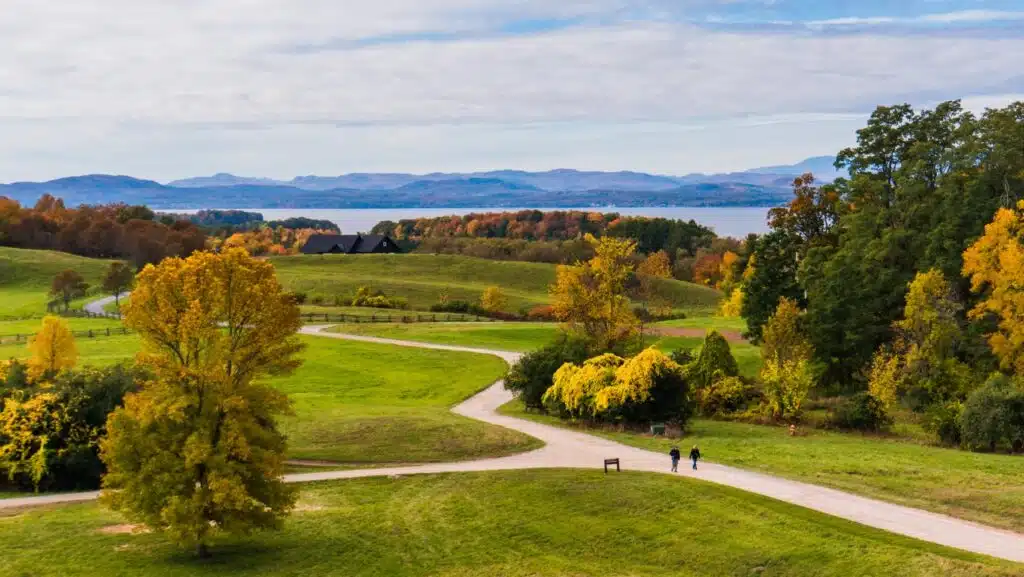 6 Scenic Spots to See New England Fall Foliage. Winding path through green hills, autumn trees, two people walking, lake and mountains in background.