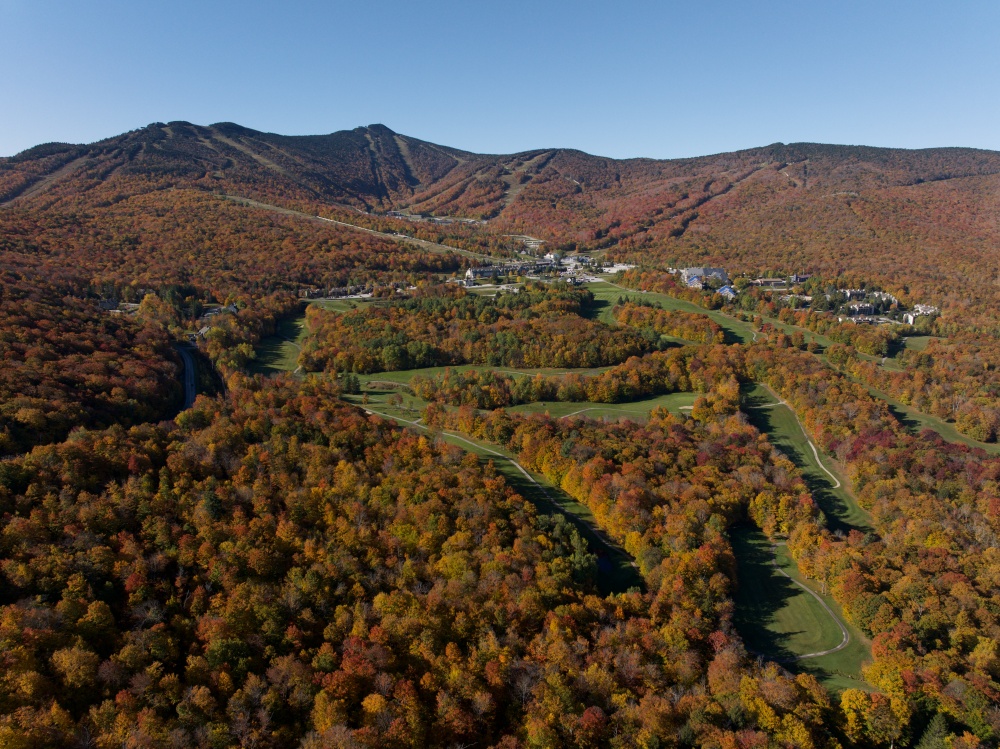 Aerial view of a golf course surrounded by autumn trees and mountains under a clear blue sky.