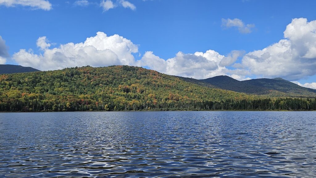 A lake with rippling water is in the foreground, bordered by a forested hillside showing early autumn colors under a partly cloudy sky.