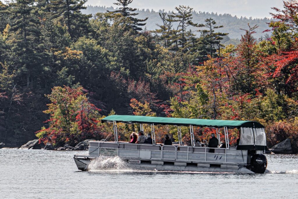 6 Peaceful Fall Foliage Destinations in New England. A small covered tour boat with several passengers moves across a lake, surrounded by trees with green and red autumn foliage.