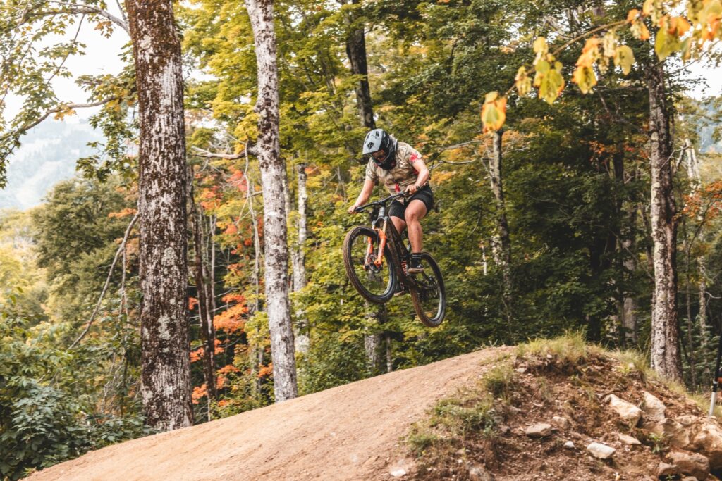 Person wearing a helmet jumps a mountain bike off a dirt trail ramp in a forest.