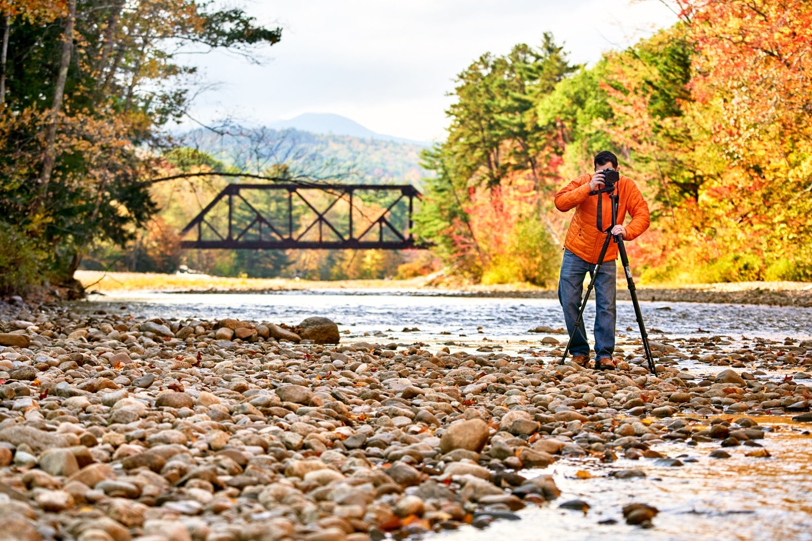 Person in an orange jacket taking a photo by a rocky river with a bridge and autumn trees in the background.
