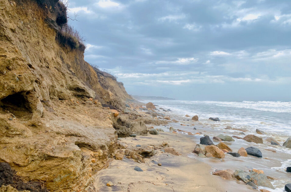 Rocky cliffs and boulders line a cloudy, windswept beach with waves crashing onto the shore.
