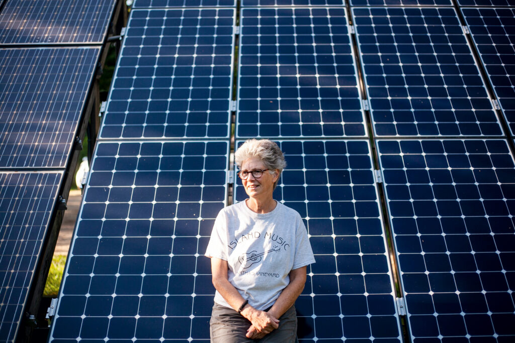 Older woman with gray hair sits smiling in front of large solar panels on a sunny day.