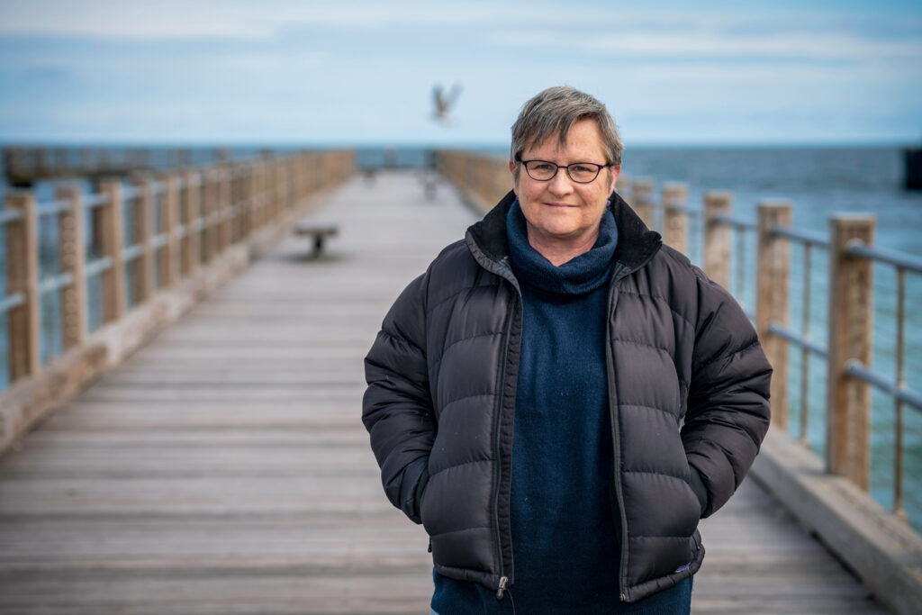 Person in a black jacket stands on a wooden pier by the sea, with a seagull in the background.