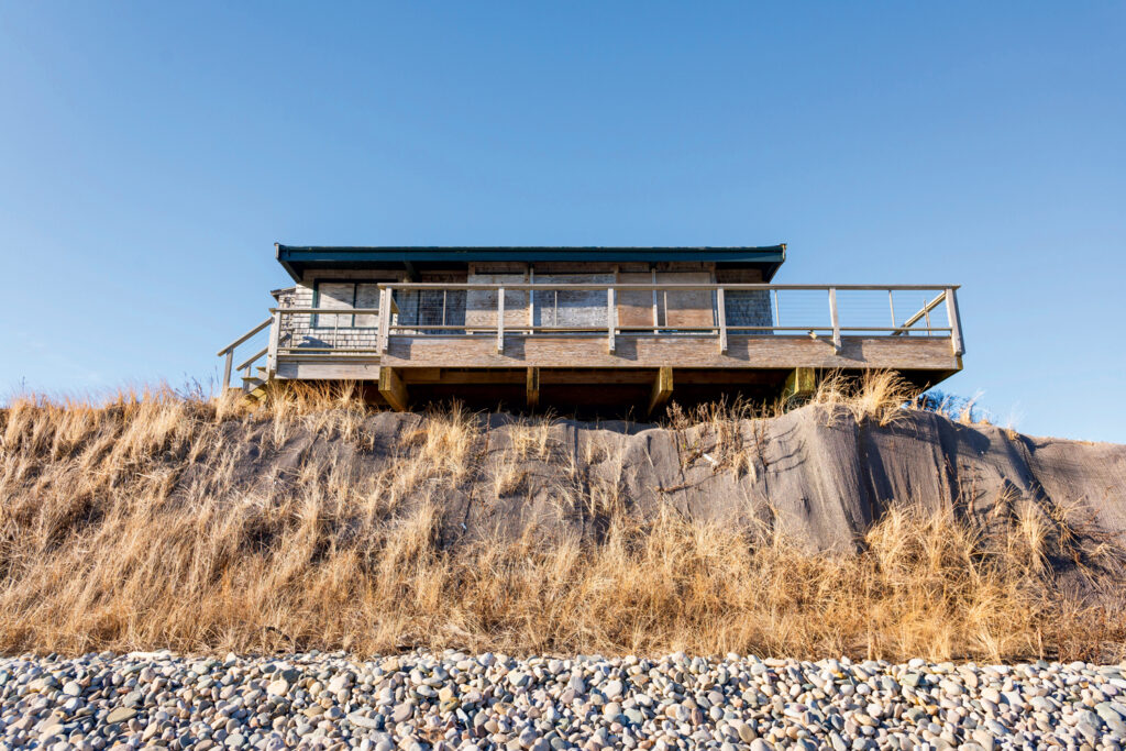 A wooden house with a deck sits atop a grassy, rocky embankment under a clear blue sky.