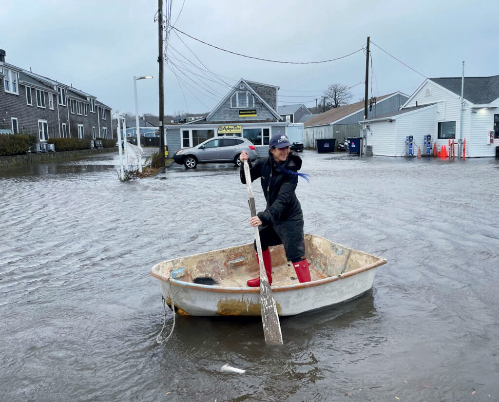 A person in red boots paddles a small boat through a flooded street in a residential area.