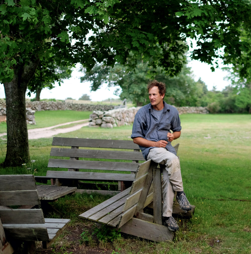A man sits on a wooden bench in a green park, holding a tool, with trees and a stone wall in the background.