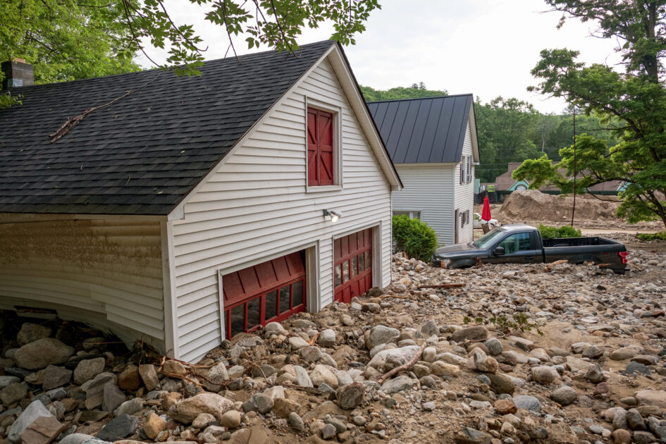 White house and truck surrounded by rocks and debris after a flood, garage doors partially buried.