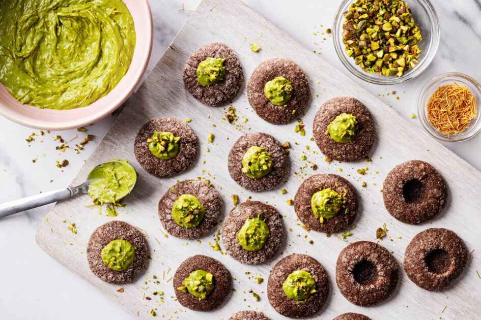Dubai Chocolate Thumbprint Cookies filled with green pistachio cream, surrounded by crushed pistachios and a bowl of frosting.