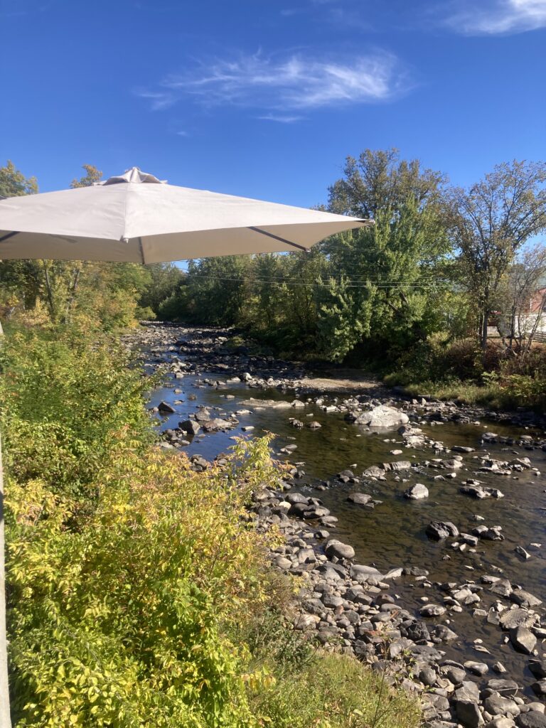 White patio umbrella over a rocky river with green trees and a clear blue sky in the background.