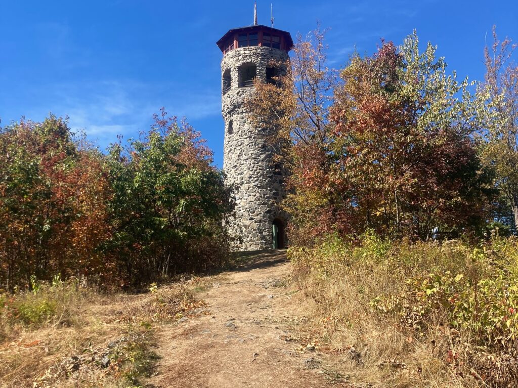Stone tower rises above a dirt path, surrounded by colorful autumn trees under a clear blue sky.
