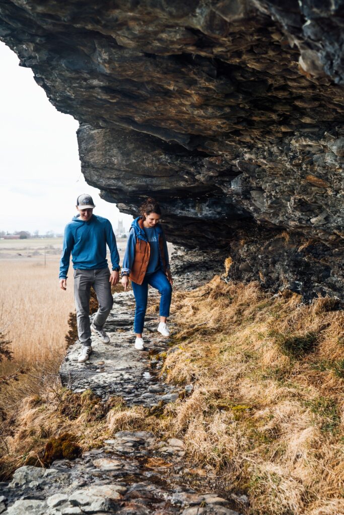 Two people walk hand in hand along a narrow rocky ledge beneath an overhanging cliff, surrounded by dry grass and an open landscape.