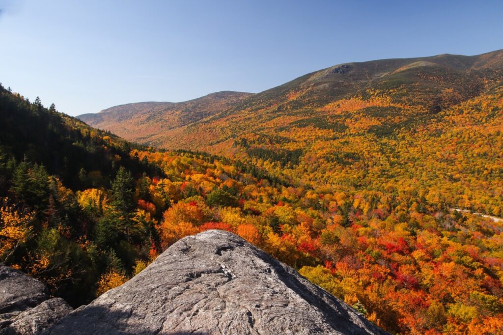 Rocky outcrop overlooks colorful autumn trees and rolling mountains under a clear blue sky.