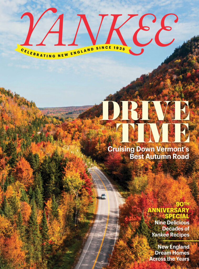 A car drives on a winding road through colorful autumn trees in Vermont under a partly cloudy sky.