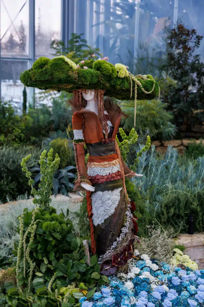 A mannequin wearing a gown and oversized hat crafted from moss, leaves, and flowers.