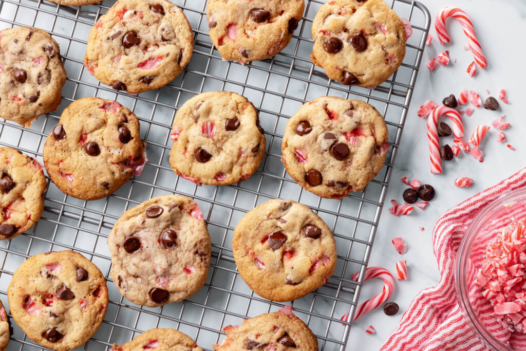 Chocolate chip and peppermint cookies cooling on a wire rack with candy canes and chocolate chips nearby.