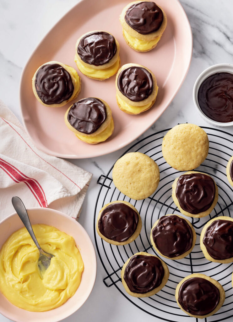 Boston cream pie cookies with chocolate glaze and yellow custard filling on plates and a cooling rack.