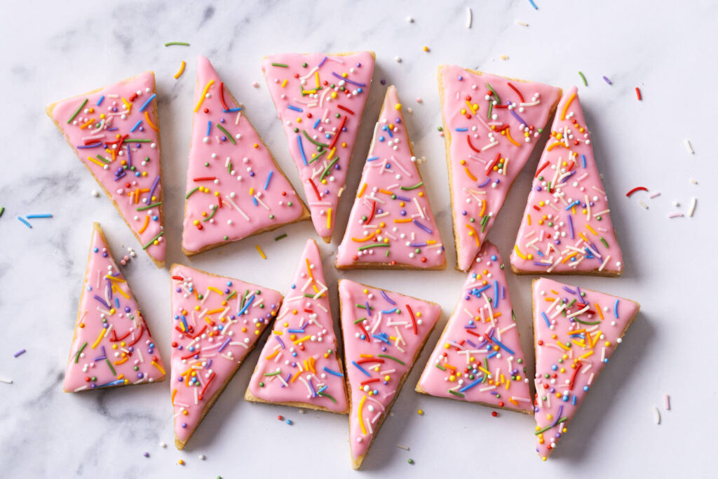 Christmas Tree Sugar Cookie Bars. Triangular sugar cookies with pink icing and colorful sprinkles arranged on a white marble surface.