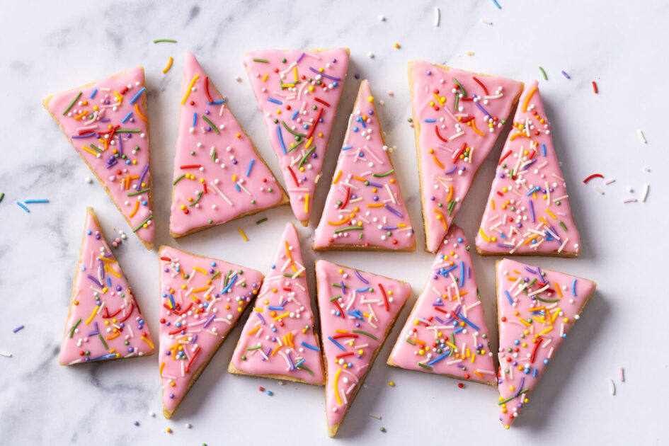Christmas Tree Sugar Cookie Bars. Triangular sugar cookies with pink icing and colorful sprinkles arranged on a white marble surface.