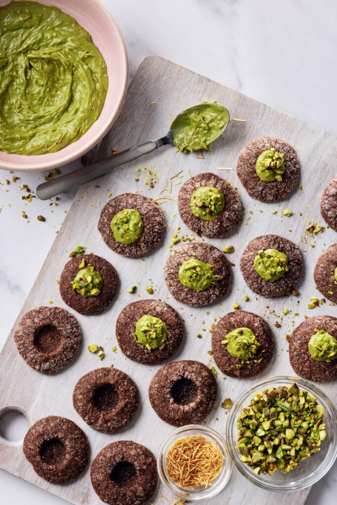 Chocolate thumbprint cookies with green pistachio filling on a board, surrounded by chopped pistachios and a bowl of filling.