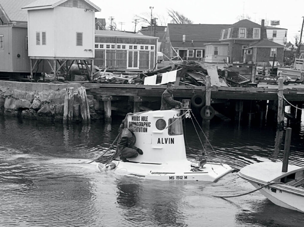 Two people on the small research submersible Alvin near a dock with damaged buildings in the background.