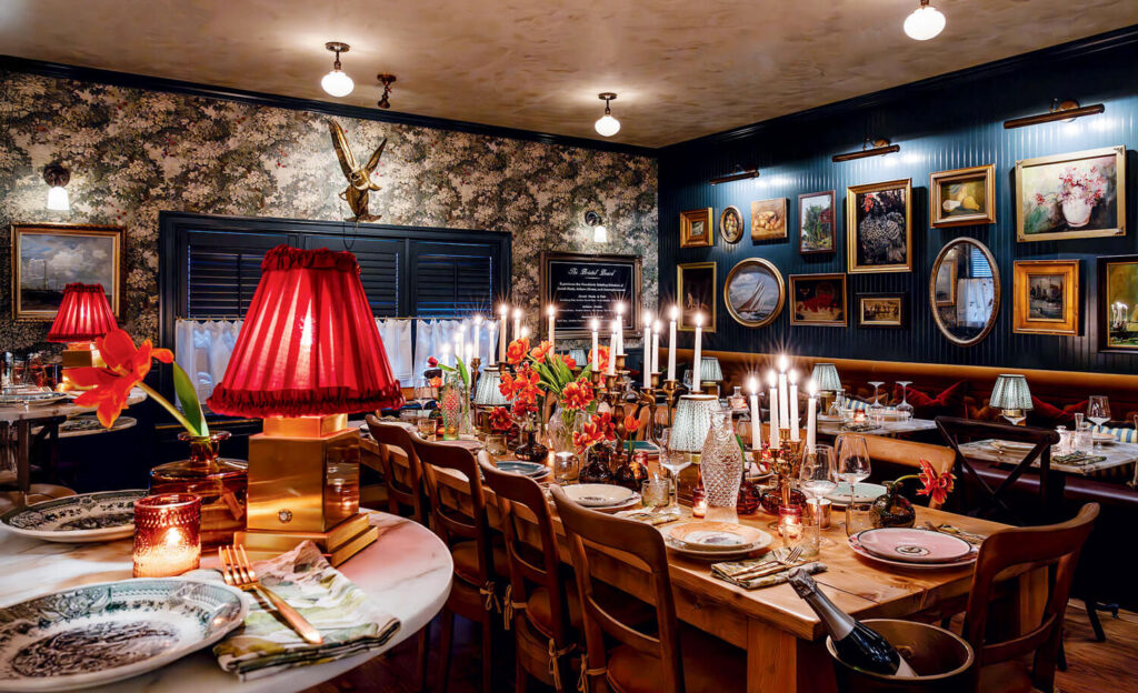 Elegant, warmly lit dining room with long tables, candles, red lamps, and vintage art on the walls.