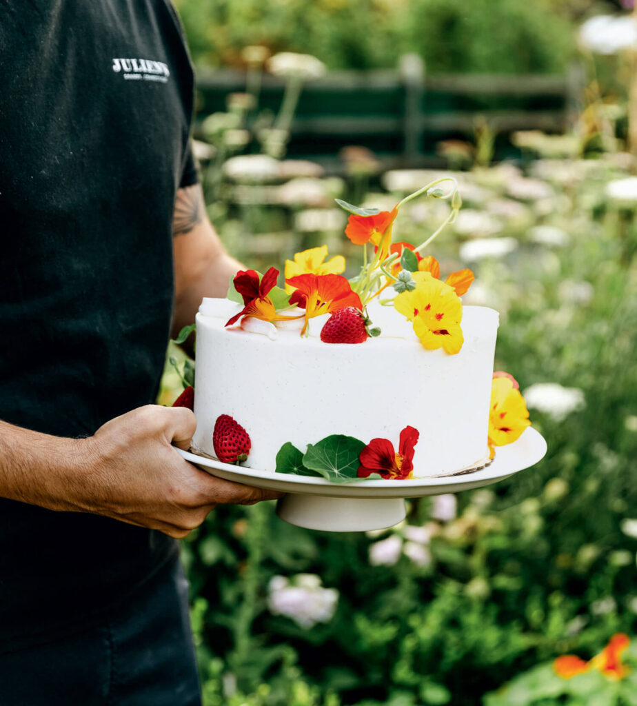 Person holding a white cake decorated with strawberries and colorful edible flowers, outdoors in a garden.