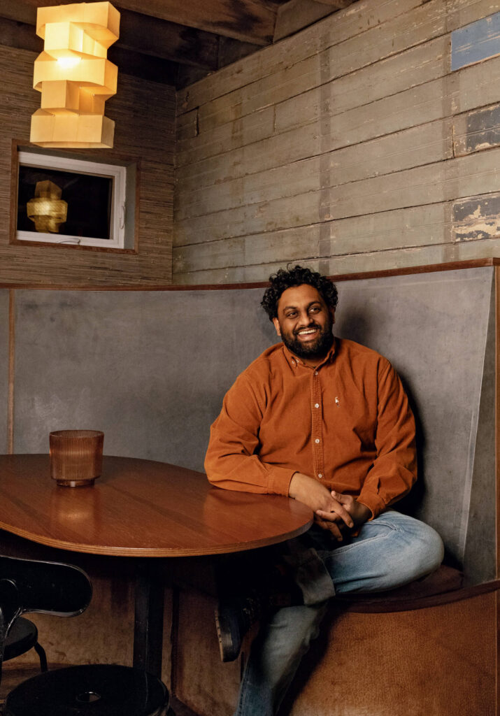 The 2025 Yankee Food Awards. A man in an orange shirt sits and smiles at a wooden table in a cozy, rustic restaurant booth.