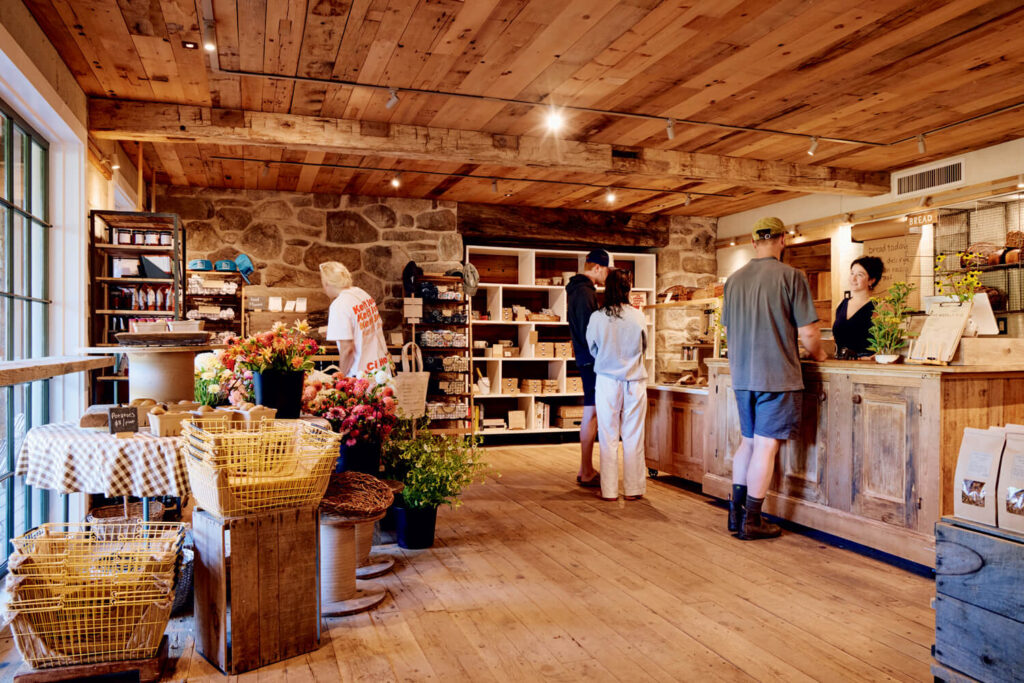 Farm shop interior with wooden décor, customers at the counter, shelves of goods, and baskets of flowers on display.